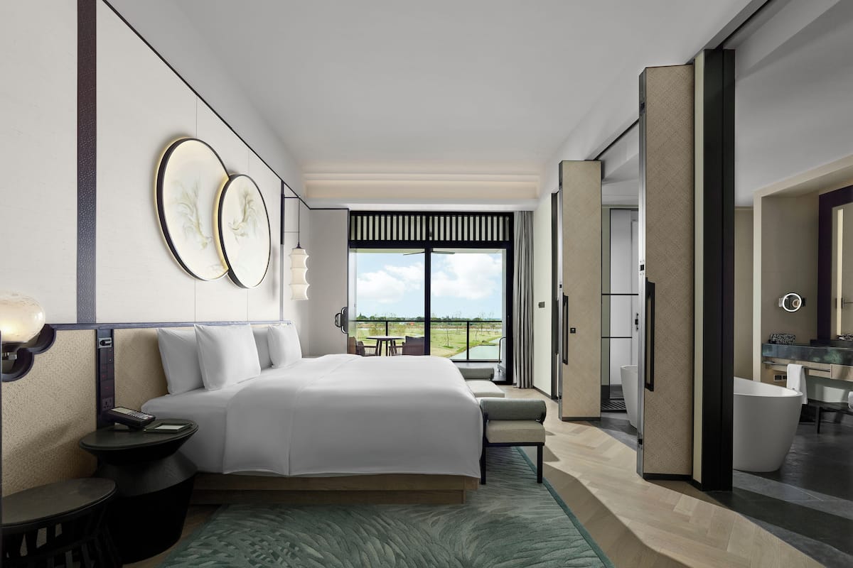 A modern hotel room with a large bed, a desk with a lamp, a view of the outdoors, and a bathroom with a bathtub.