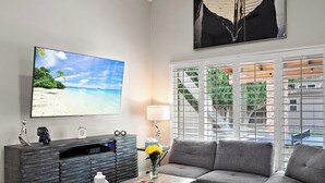 Ein Wohnzimmer mit großem Flachbildfernseher, einem grauen Sofa, einem Couchtisch mit einer Vase voller gelber Blumen und einer Vitrine mit Dekorationsgegenständen.