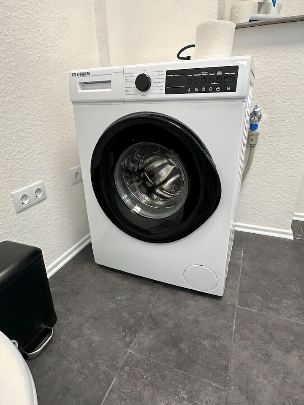 A front-loading washing machine in a laundry room with a white wall and gray floor tiles.