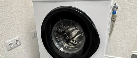 A front-loading washing machine in a laundry room with a white wall and gray floor tiles.