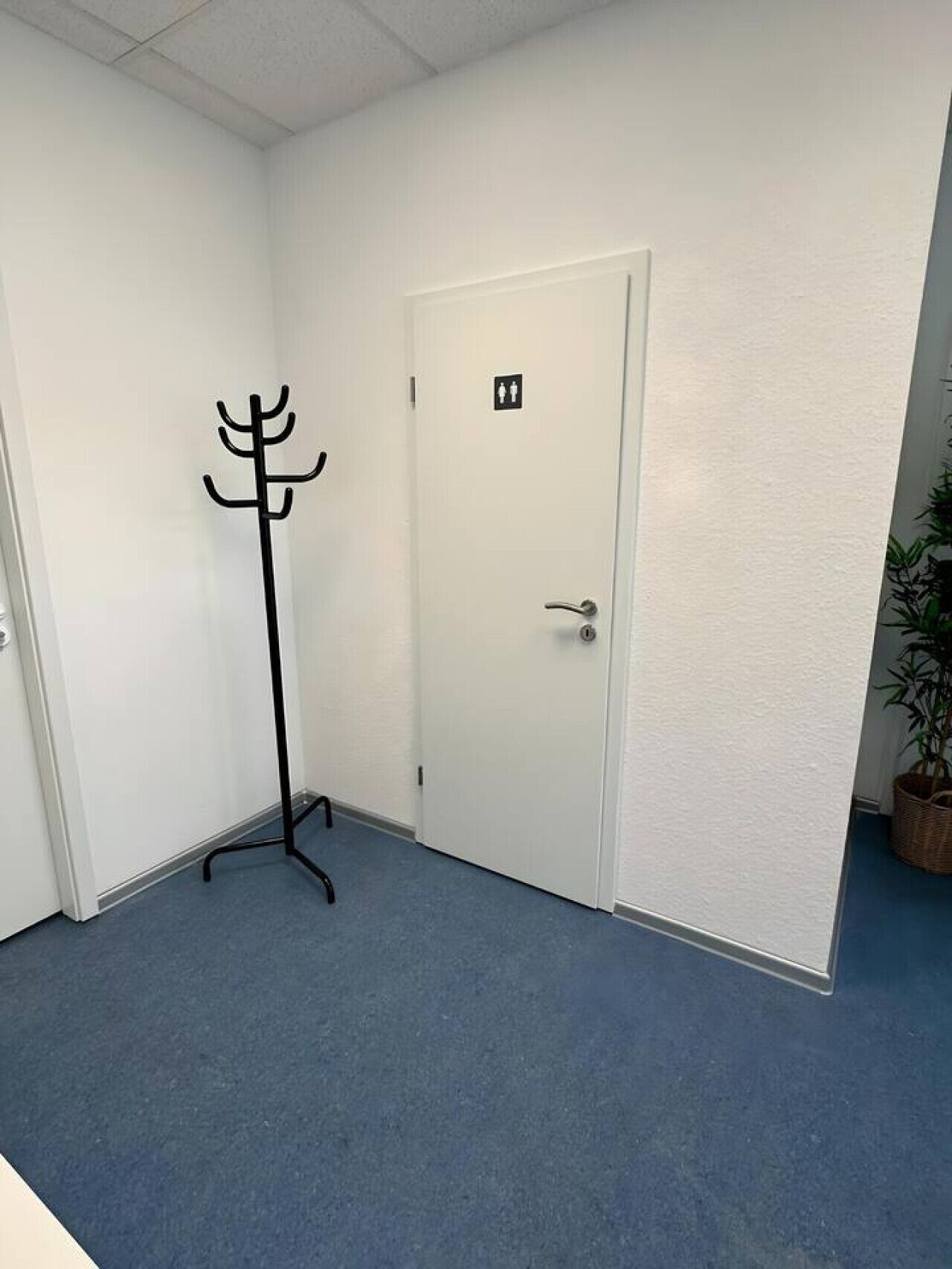 Ein Zimmer mit Kleiderständer, einer Tür mit Schildern für Damen- und Herrentoilette sowie einem Blumentopf.
