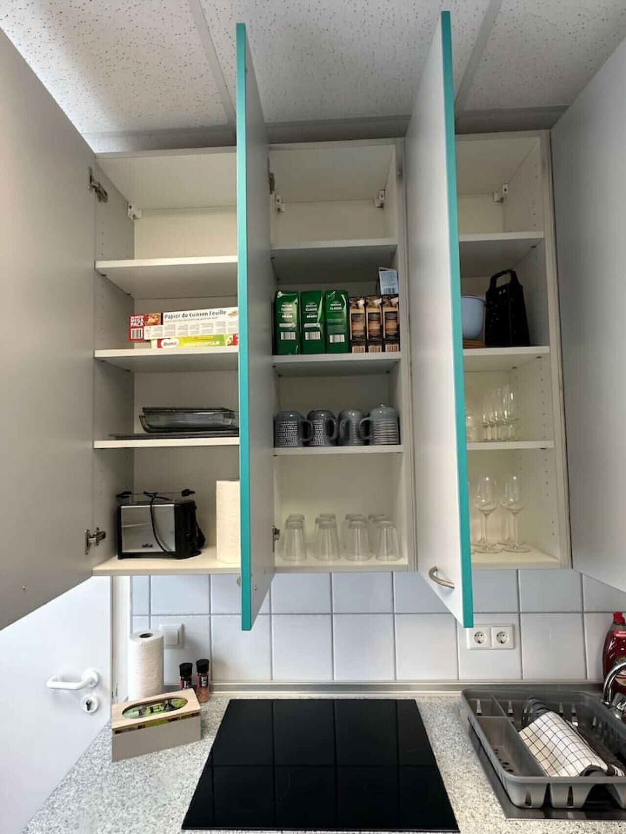 Eine Küche mit weißen Schränken, einer schwarzen Arbeitsplatte und einer türkisblauen Spritzschutzwand. Es sind Regale mit verschiedenen Gegenständen zu sehen, darunter ein Toaster, eine Kaffeemaschine und Gläser.