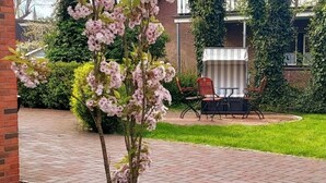 A patio area with a brick pathway, a small tree with pink flowers, and outdoor furniture. - Holiday Apartment Waterdelle (Borkum)