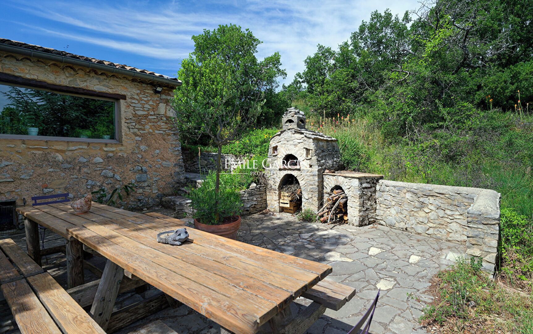 A stone building with a wooden table and benches, surrounded by greenery and a stone wall.