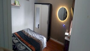 Un dormitorio con una cama, un armario, un espejo con luz y estantes con objetos.