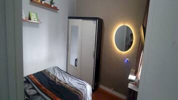 A bedroom with a bed, a wardrobe, a mirror with a light, and shelves with items.