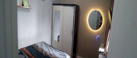 A bedroom with a bed, a wardrobe, a mirror with a light, and shelves with items.