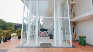 Una entrada de edificio moderna con fachada de vidrio, un contenedor verde para basura y piso de baldosas.
