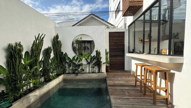 A modern outdoor pool area with a wooden deck, surrounded by lush greenery and a white building with large windows.