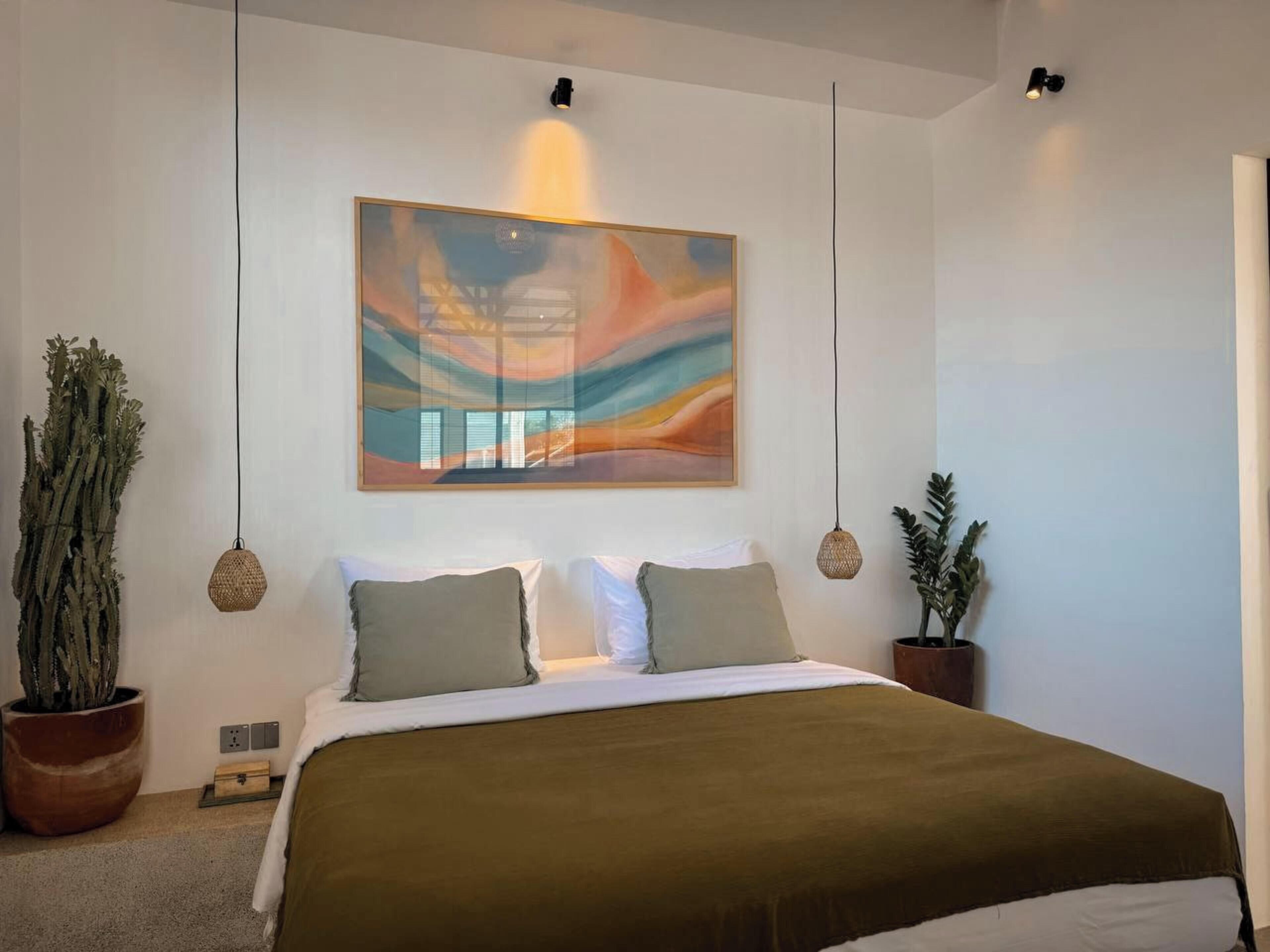 A modern bedroom with a large bed, two bedside tables, a wall-mounted abstract painting, and two potted plants.