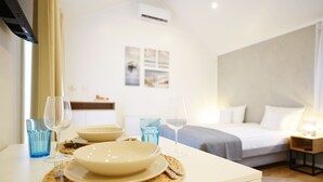 Een hotelkamer met een eethoek, waar een witte tafel staat gedekt met borden, glazen en bestek. In de achtergrond is een bed met een grijze deken en kussens te zien.