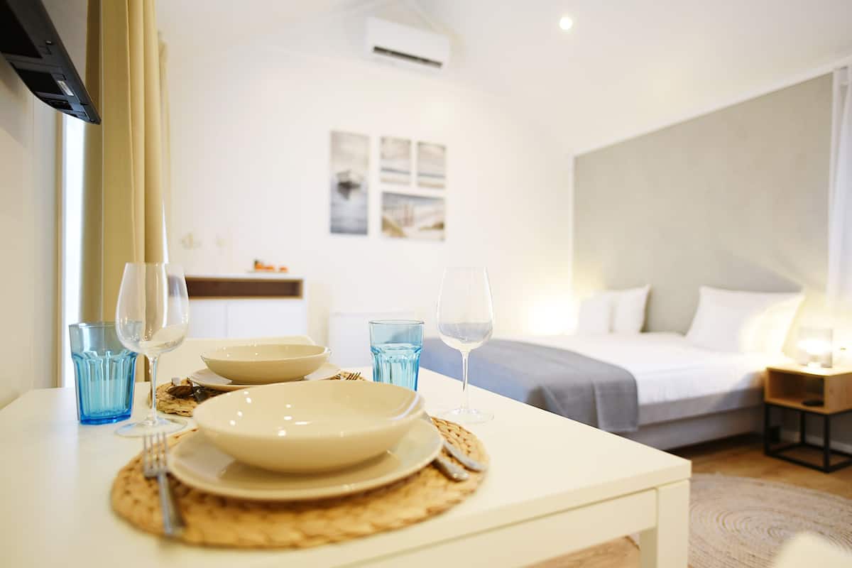 Ein Hotelzimmer mit Essbereich, inklusive weißem Tisch, gedecktem mit Tellern, Gläsern und Besteck. Im Hintergrund ist ein Bett mit grauer Decke und Kissen zu sehen.