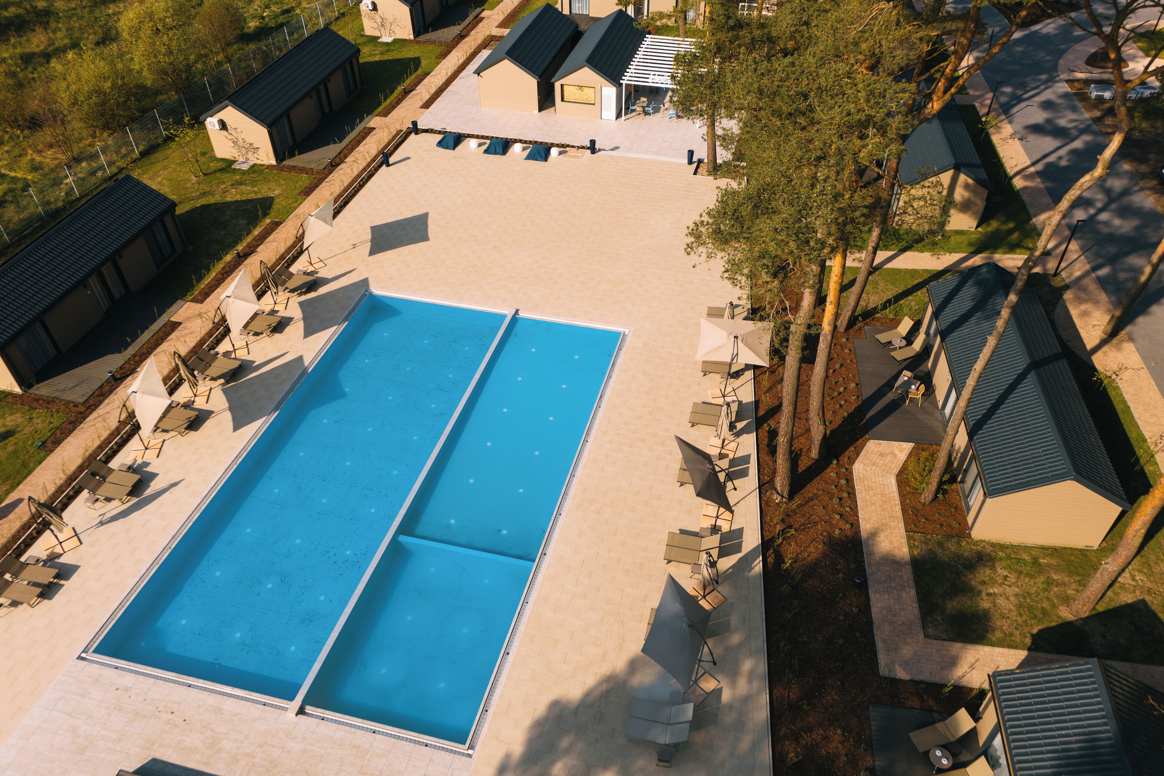 Ein großer rechteckiger Pool mit zwei separaten Bereichen, umgeben von Liegestühlen und Sonnenschirmen, mit angrenzenden Gebäuden und einem Baum im Hintergrund.