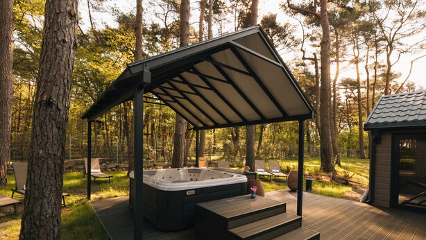 Een hottub onder een houten pergola op een terras, omringd door bomen.