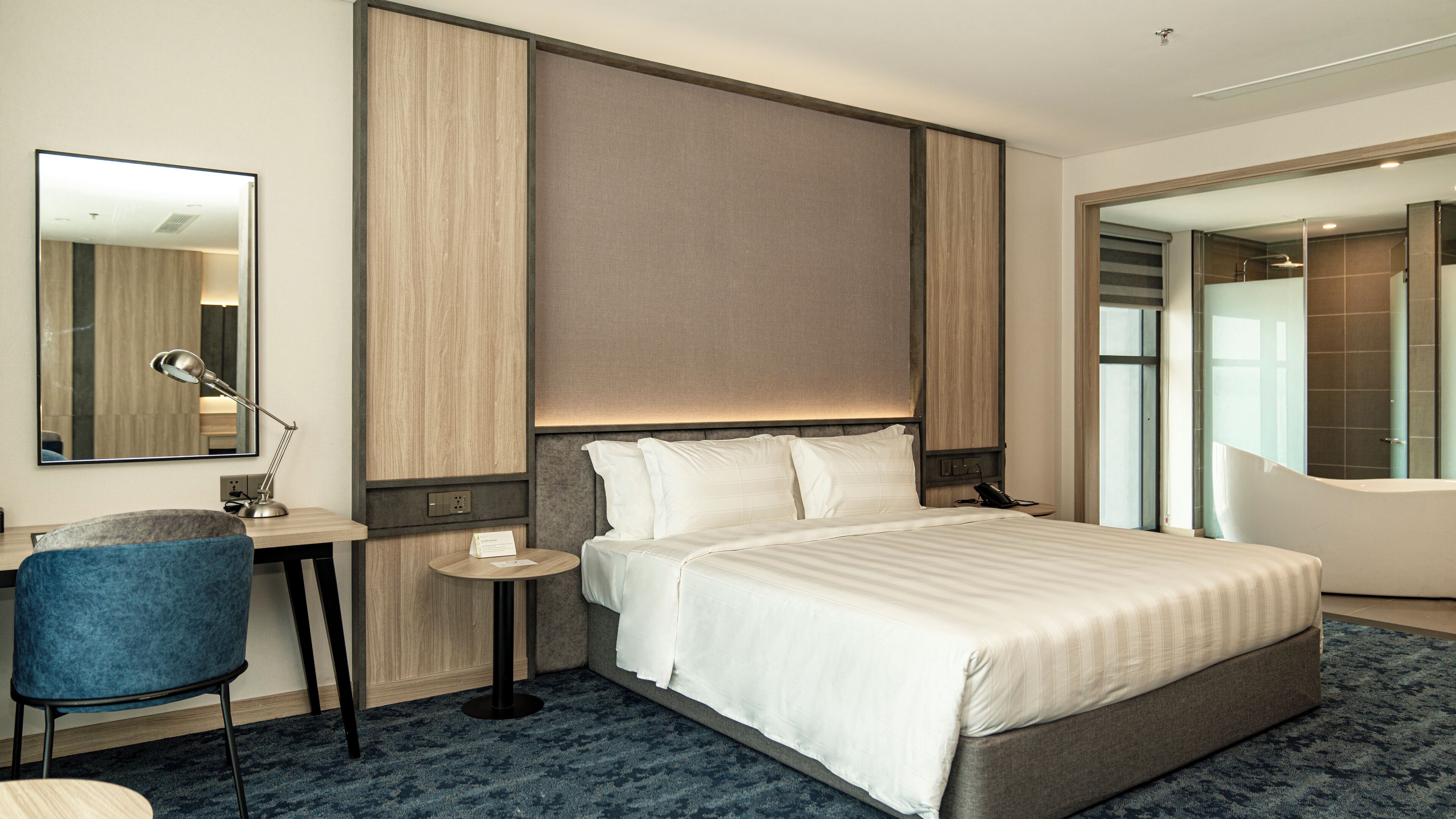 A modern hotel room with a large bed, a desk with a chair, a round side table, and a large mirror.