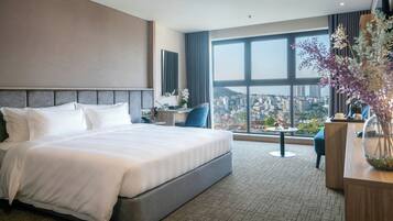 A modern hotel room with a large bed, a desk, a chair, and a view of a cityscape through large windows.
