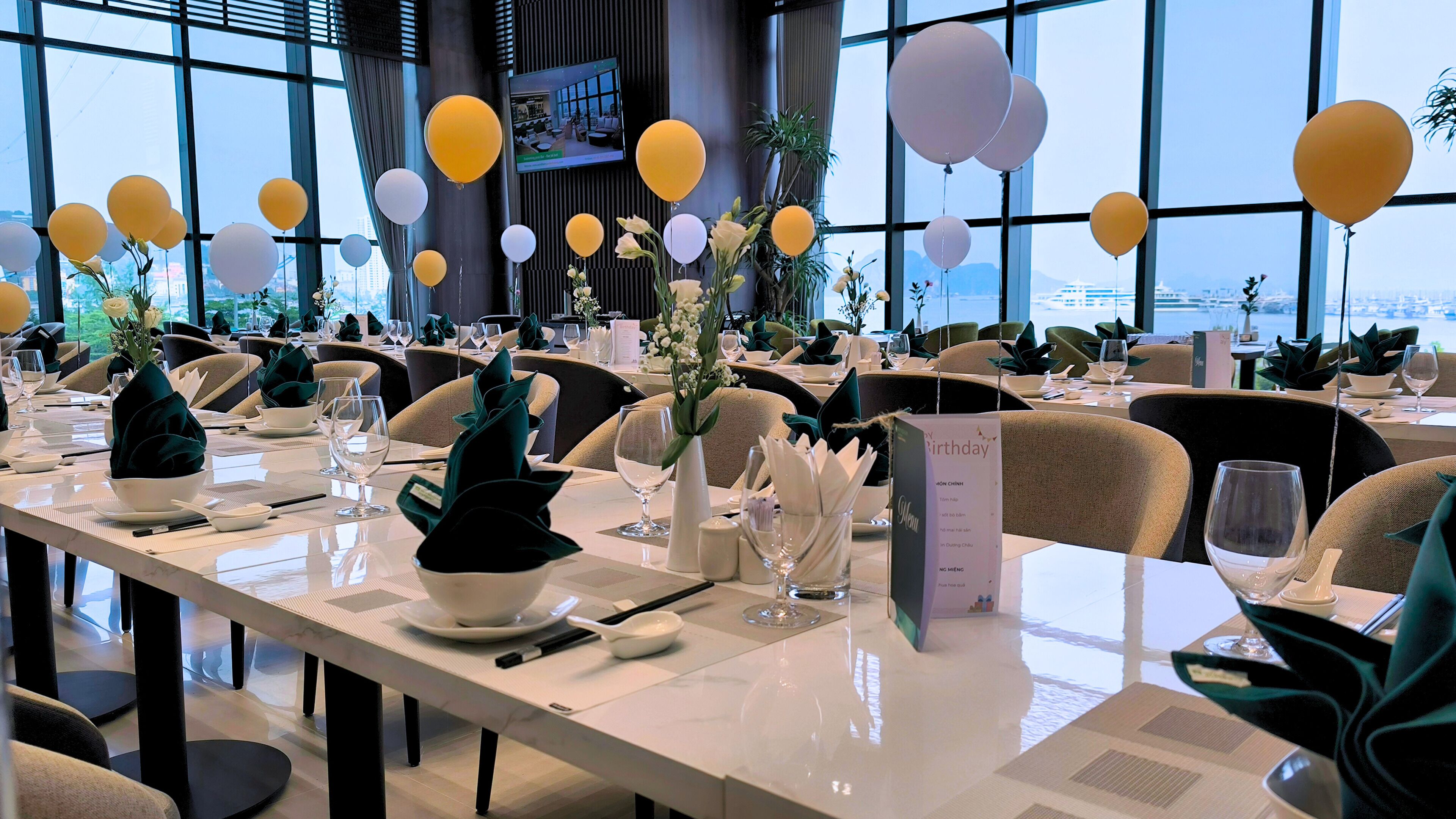 Un comedor con mesas dispuestas para un evento formal, que incluye un menú y vistas de la ciudad a través de amplios ventanales.