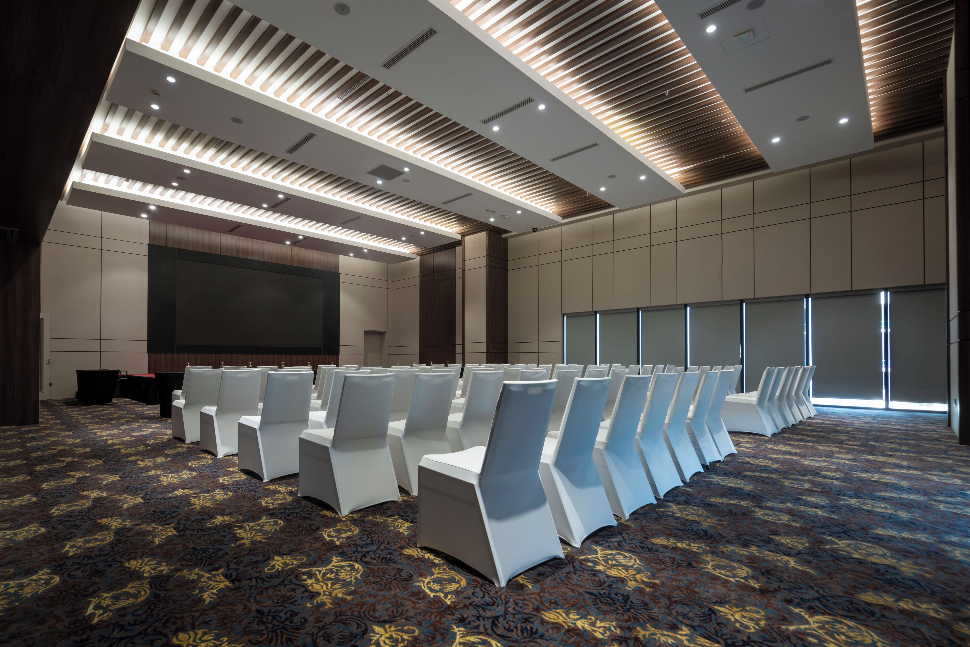 Un salón de conferencias con hileras de sillas blancas, una pantalla grande y una alfombra estampada.