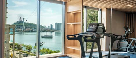 Un gimnasio con cintas de correr y equipo de ejercicio, amplias ventanas con vistas a un río y al paisaje urbano, y techo de madera.