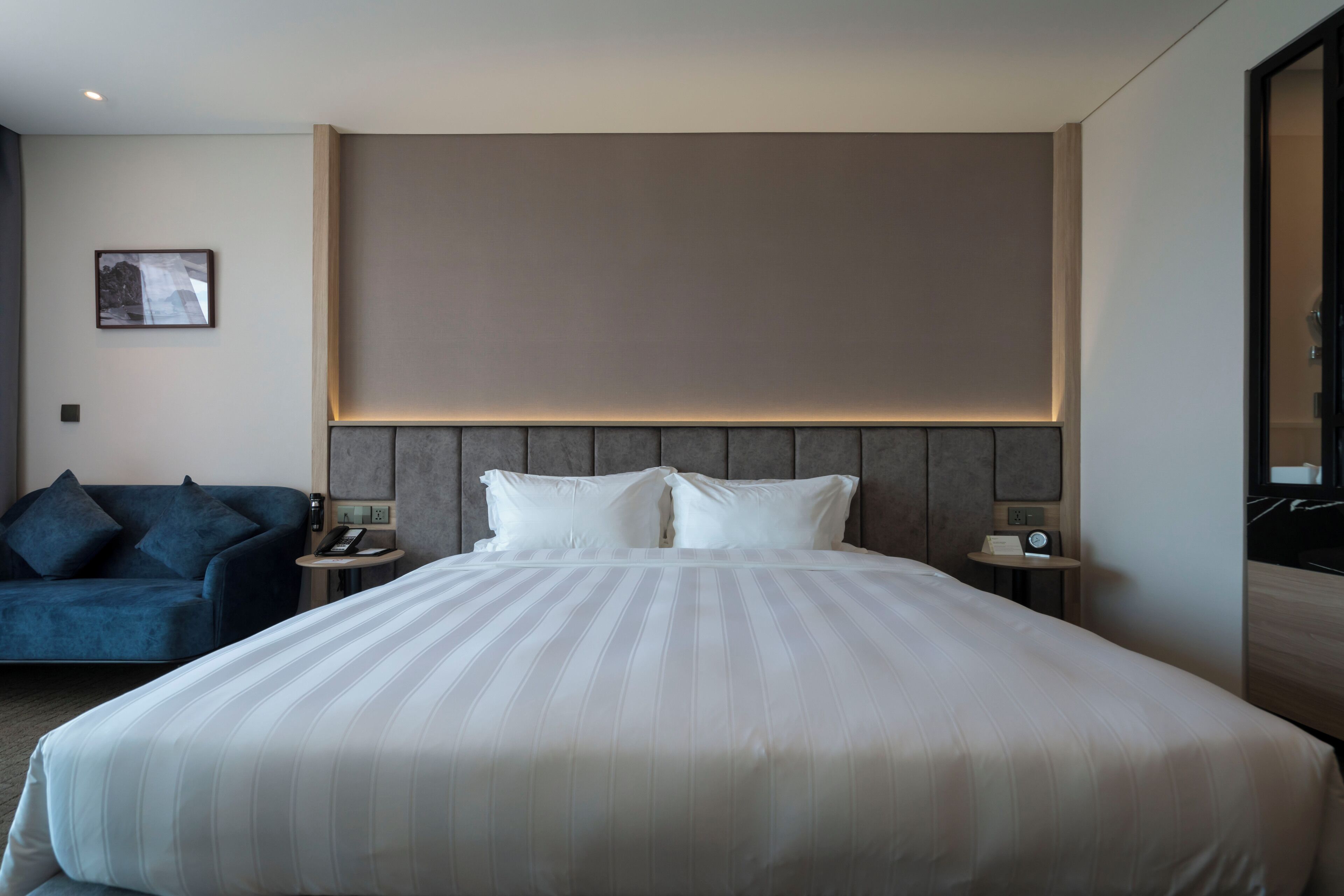 Una habitación de hotel moderna con una cama grande, un sofá azul, una mesita de noche con una lámpara y un cuadro enmarcado en la pared.