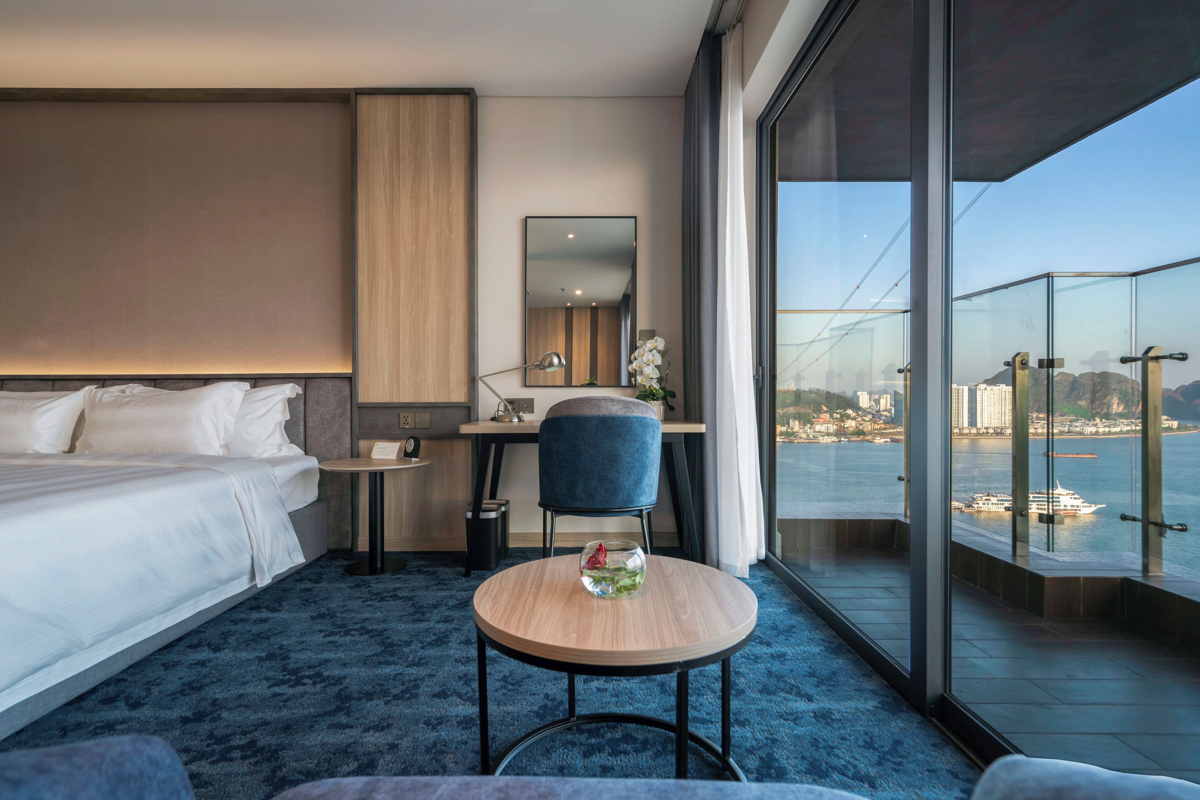 A modern hotel room with a bed, desk, chair, and a round table with a vase. A large window offers a view of a city and water.
