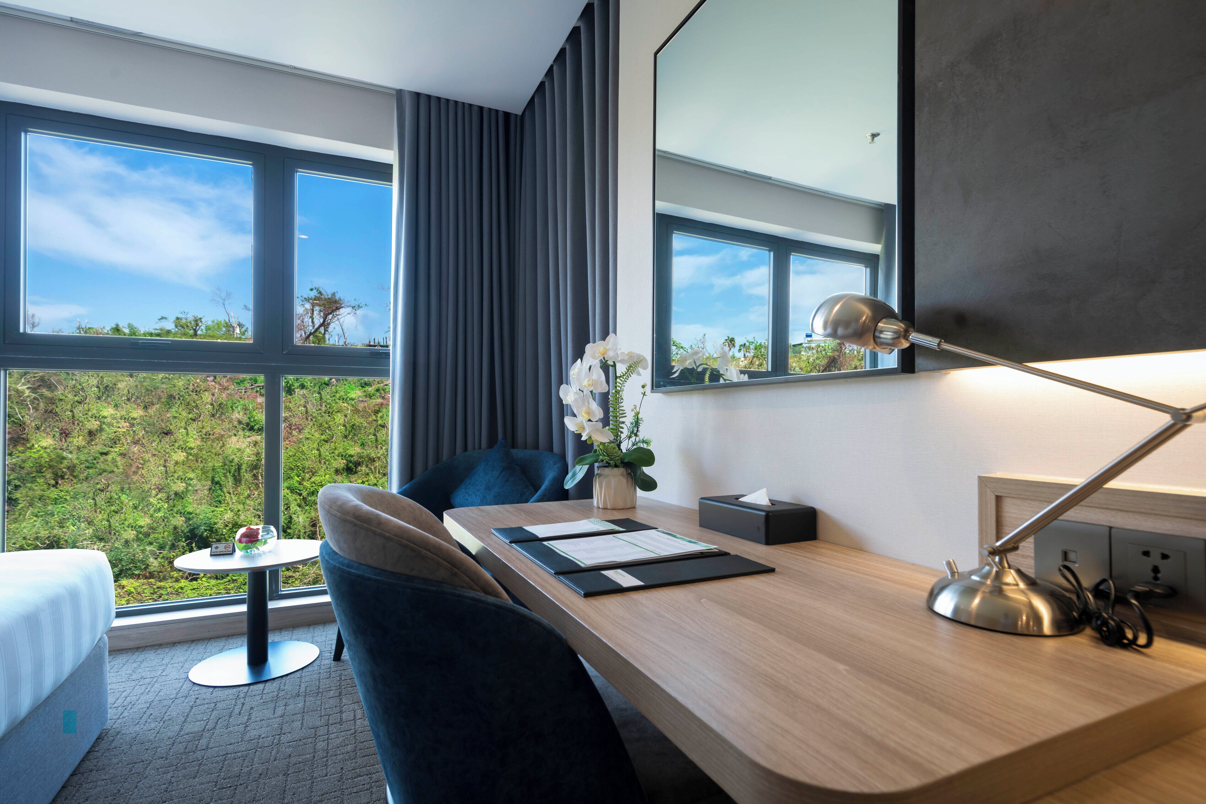 A modern hotel room with a wooden desk, a comfortable chair, a small round table, a bedside lamp, and a large window offering a view of greenery.