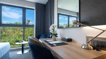A modern hotel room with a wooden desk, a comfortable chair, a small round table, a bedside lamp, and a large window offering a view of greenery.