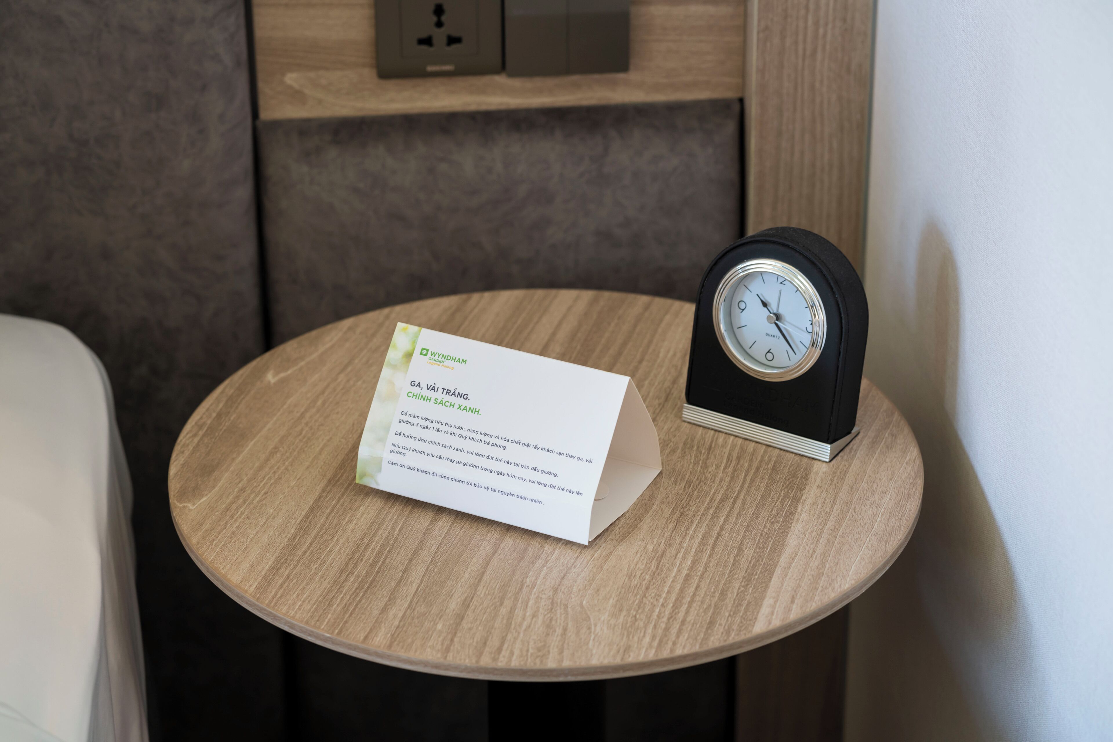 A round wooden table with a black alarm clock and a white card with text.