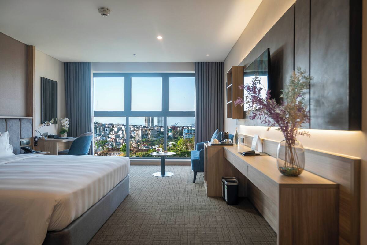 A modern hotel room with a large bed, a desk, a chair, a small table, a trash bin, a vase with flowers, a large window with a city view, and a mounted TV.