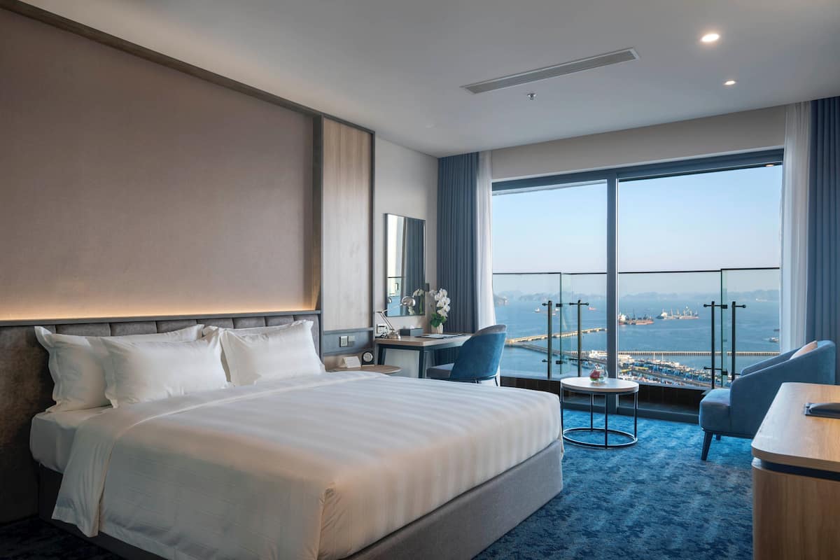 A hotel room with a large bed, a desk, a chair, and a view of the sea through large windows.