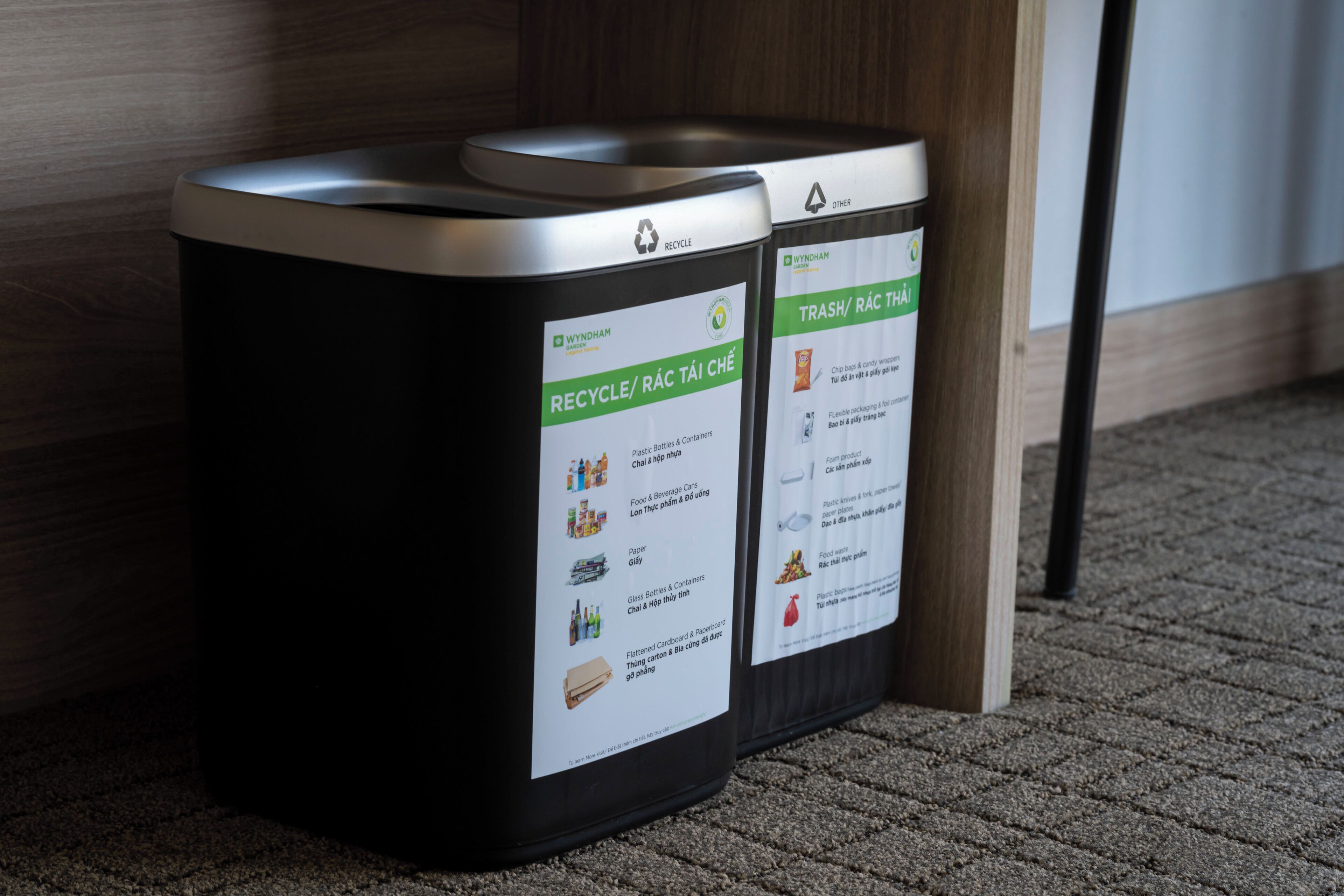 Two black trash bins with recycling and trash labels, placed on a carpeted floor next to a wooden structure.