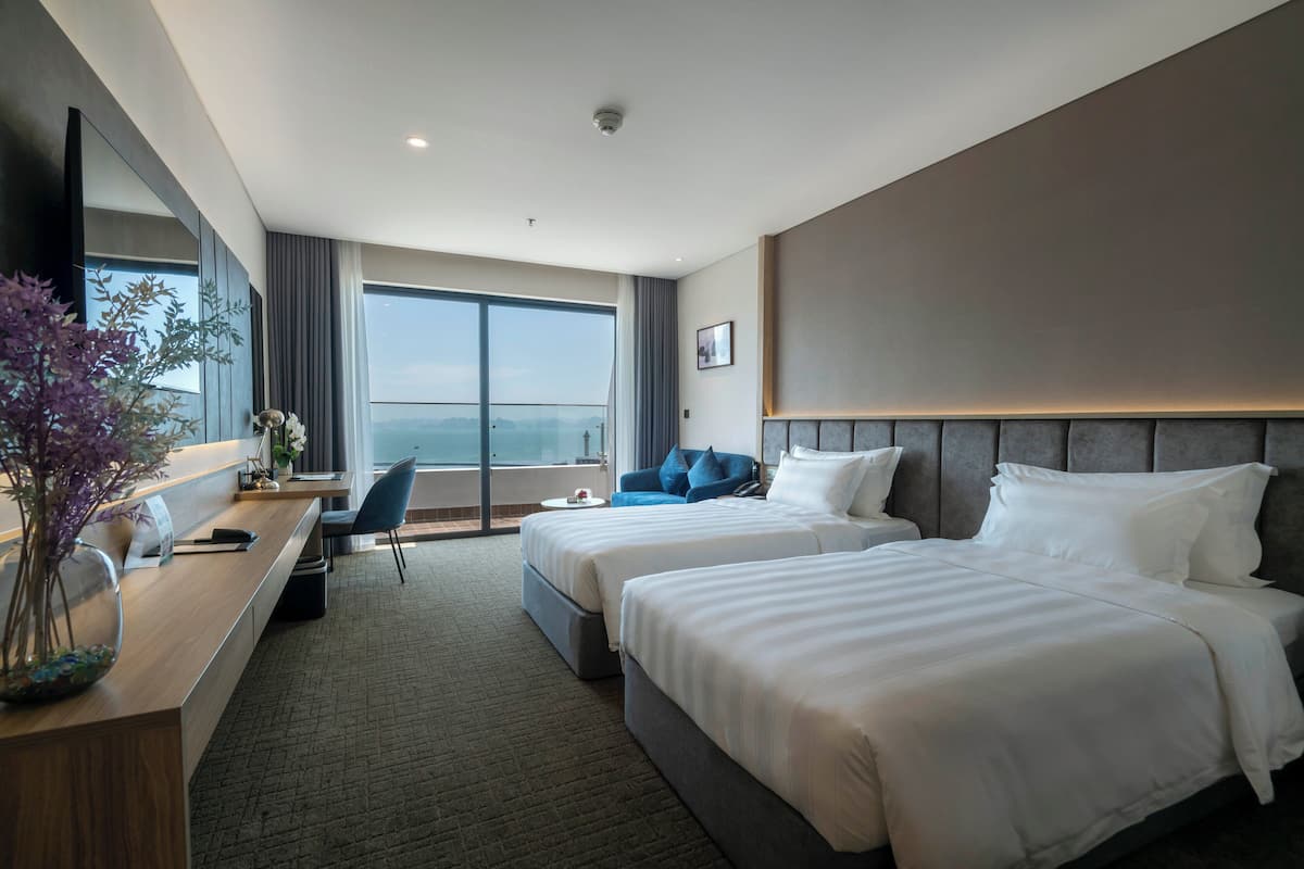 A hotel room with two beds, a desk, a chair, a large window, and a view of the city.