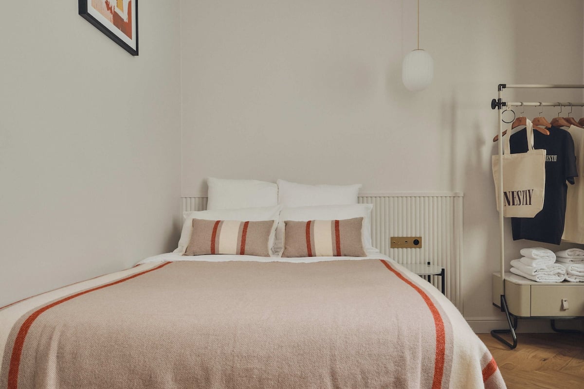A neatly made bed with a striped blanket, a bedside table with a lamp, and a clothing rack with hangers and a tote bag.