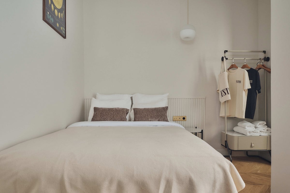 A neatly arranged bedroom with a bed, a radiator, a hanging light, a wardrobe with clothes, and a towel rack.