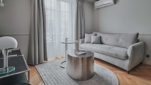 Un salon moderne avec un canapé gris, une table basse ronde en métal, une table d’appoint en verre et une grande fenêtre ornée de rideaux légers.