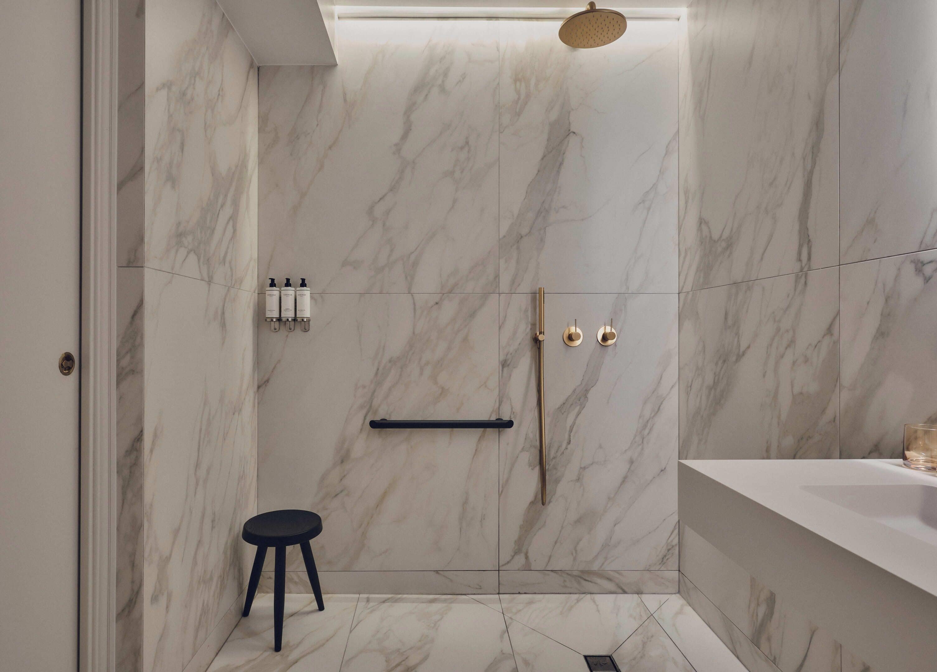 A modern bathroom with marble walls, a shower with a gold showerhead, a black stool, and a white sink.