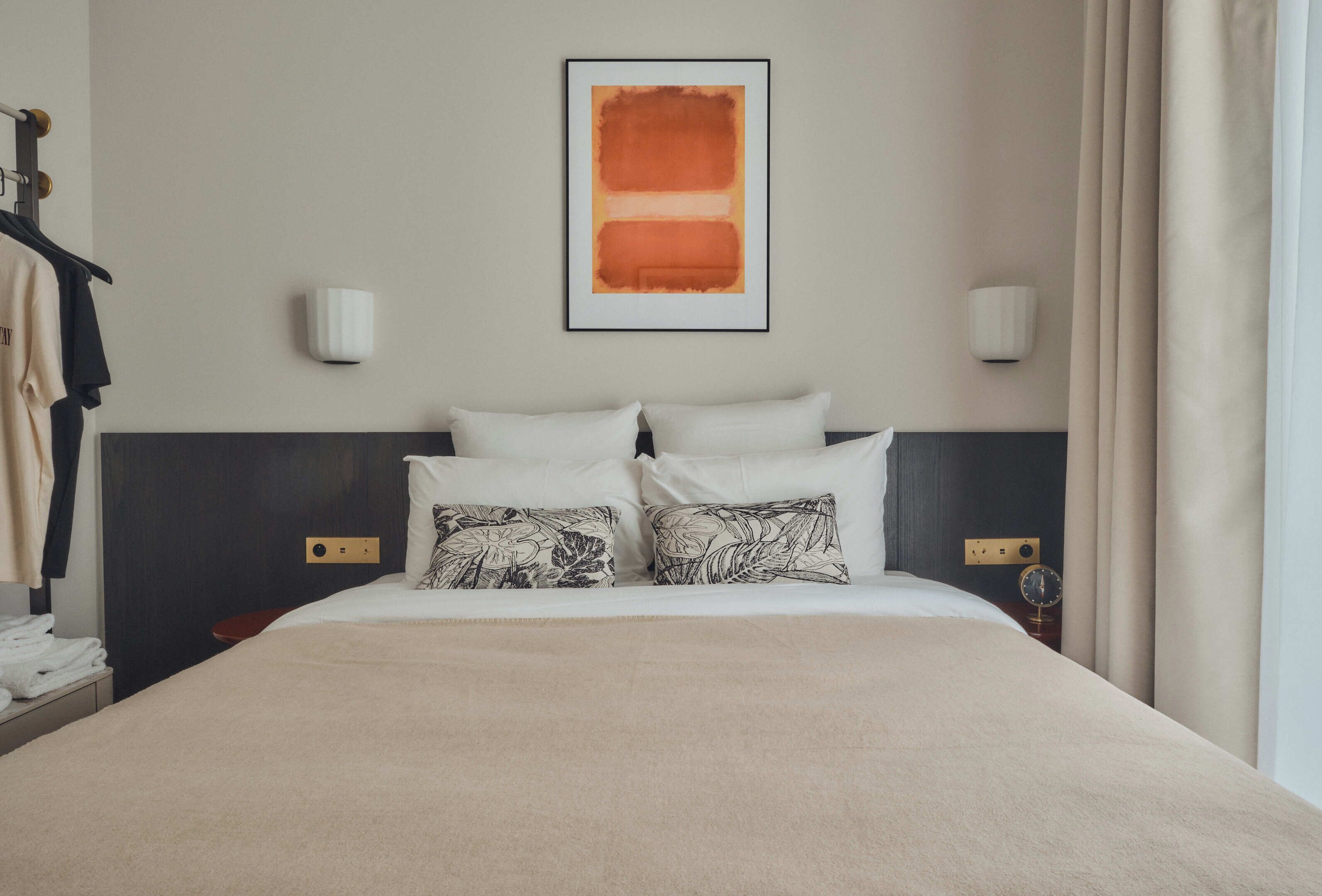 A hotel room with a large bed, two wall-mounted light fixtures, a framed artwork above the bed, and a window with curtains.