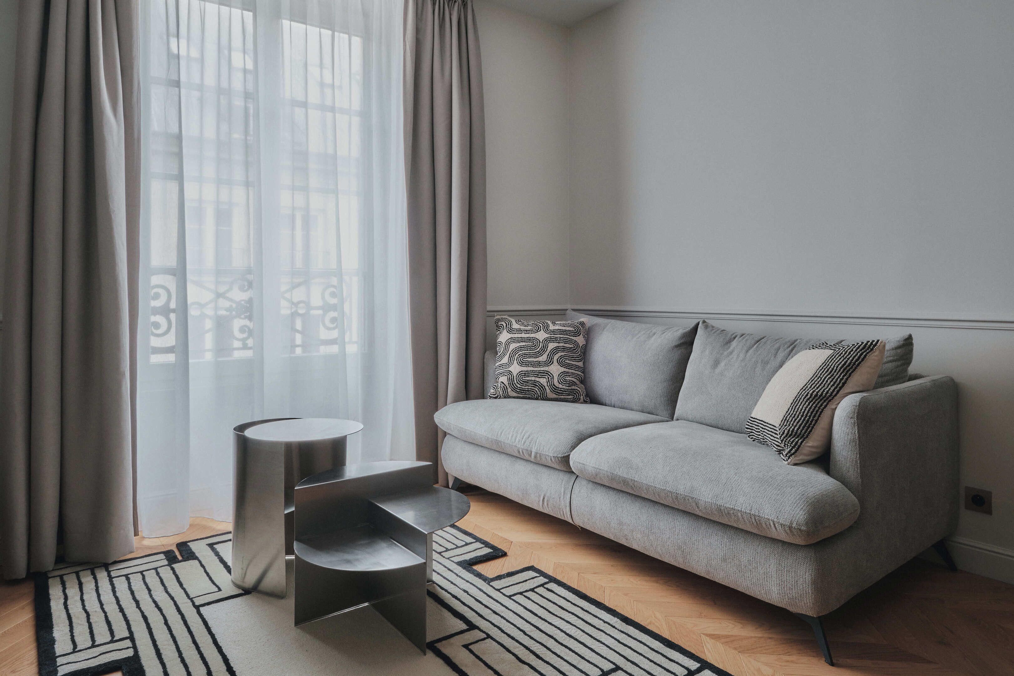 Ein modernes Wohnzimmer mit einem grauen Sofa, gemusterten Kissen, einem kleinen Beistelltisch und einem geometrischen Teppich.
