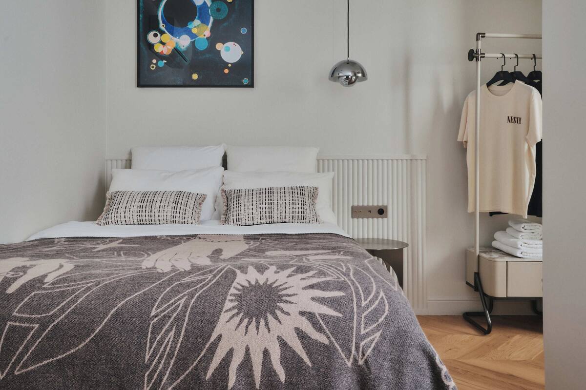 A modern bedroom with a bed, a wall-mounted abstract painting, a hanging pendant light, a radiator, a clothing rack with a shirt, and a folded towel on a bedside table.