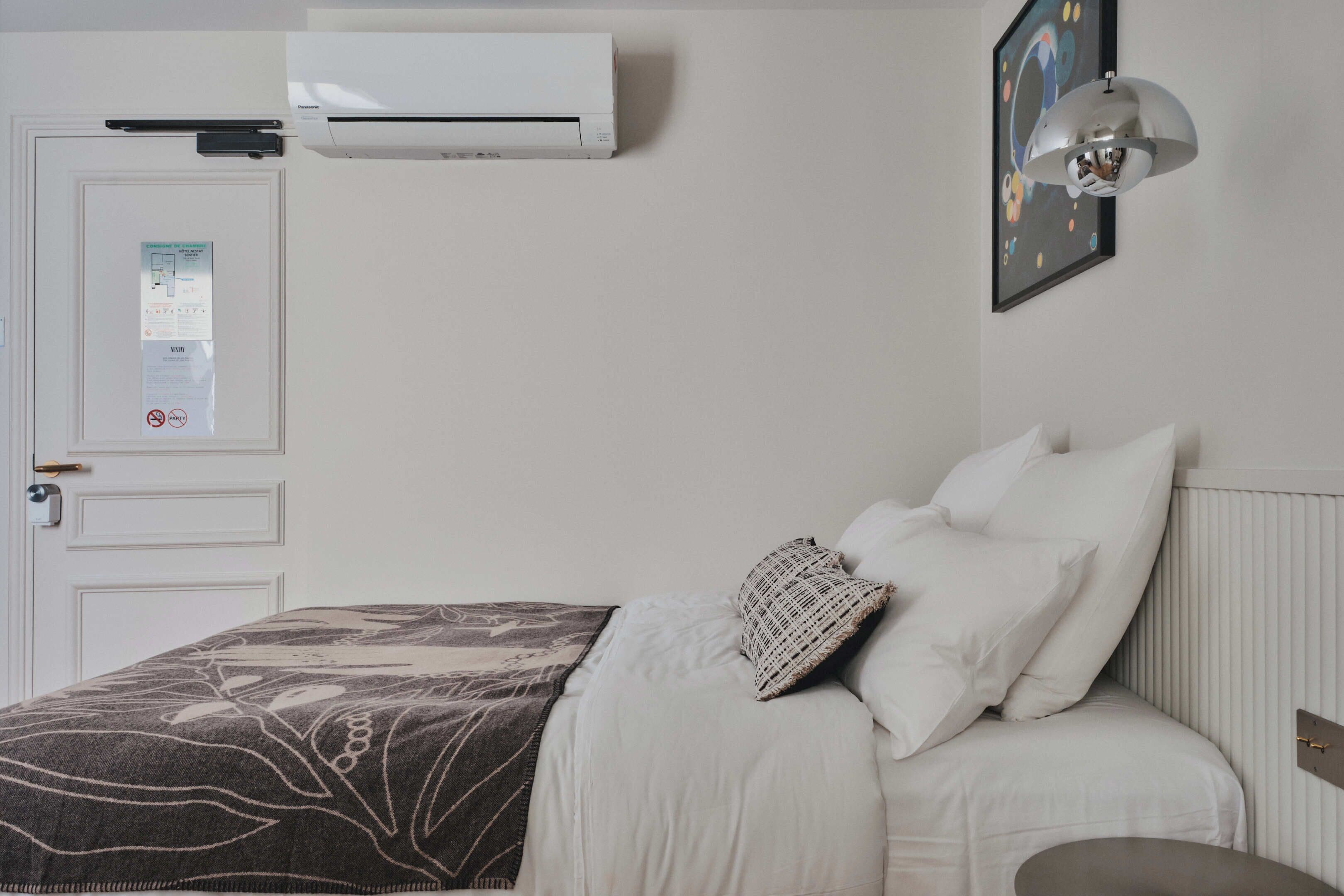 Un dormitorio con cama, ropa de cama blanca, aire acondicionado de pared, una puerta con un aviso y un cuadro en la pared.