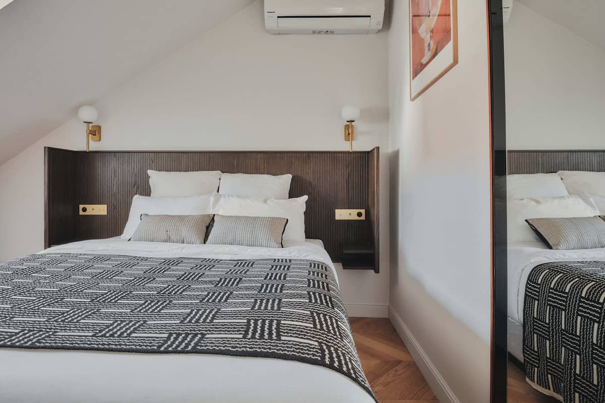 A neatly made bed with a patterned bedspread, a wooden headboard, bedside lamps, and a wall-mounted air conditioner.