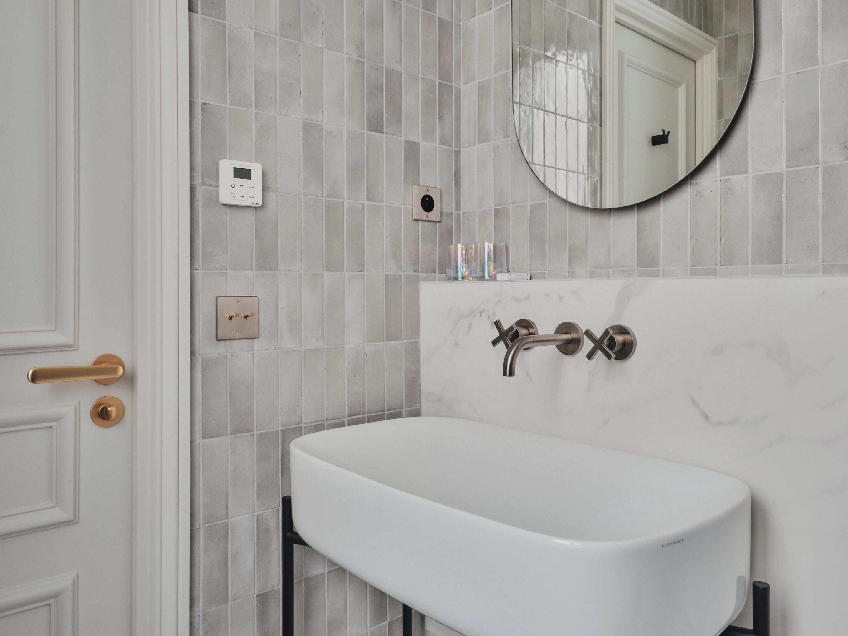 Une salle de bain avec un lavabo blanc, un miroir rond et une porte dotée d’une poignée dorée.