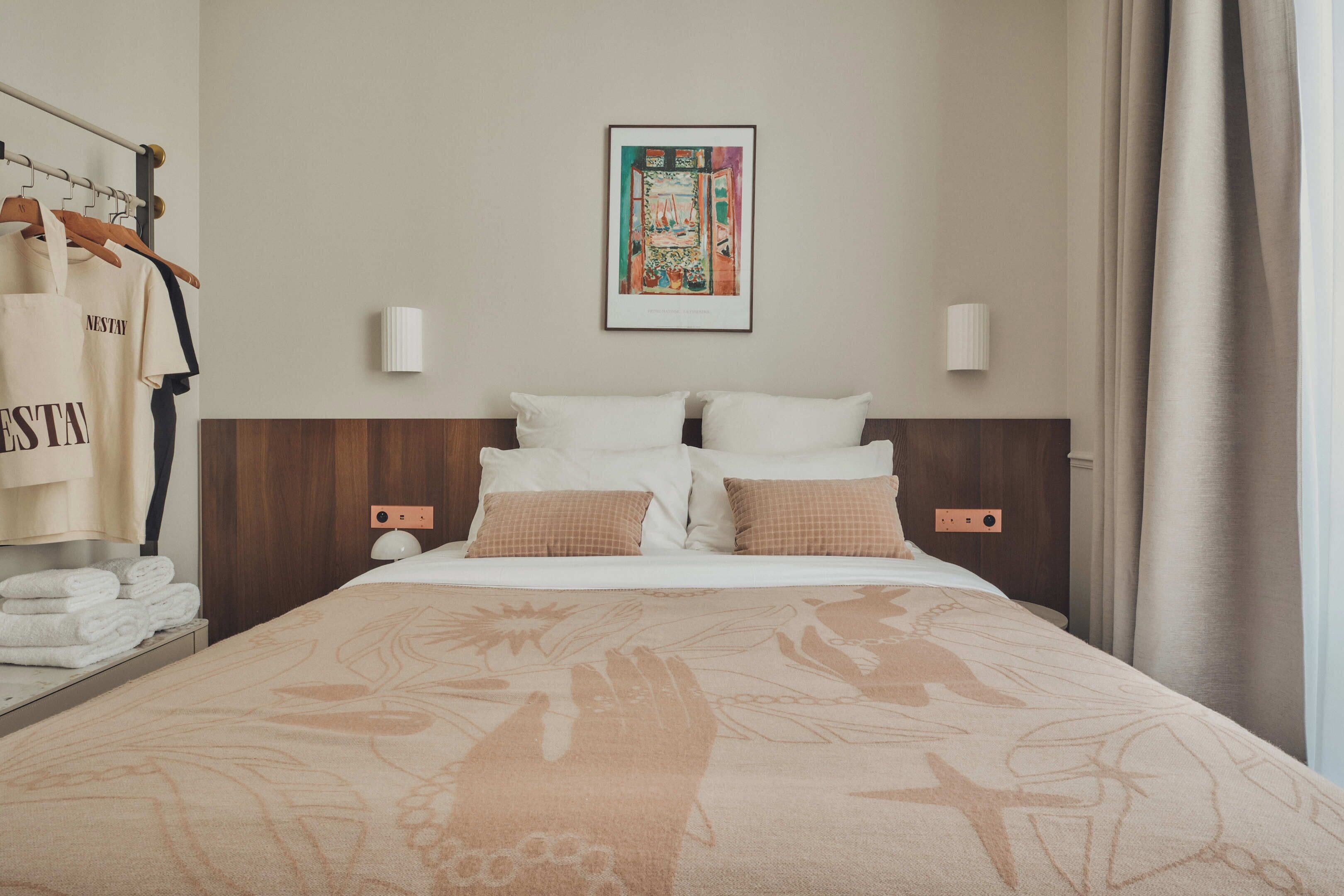 A hotel room with a bed, pillows, a framed artwork on the wall, a wardrobe, and a towel rack.