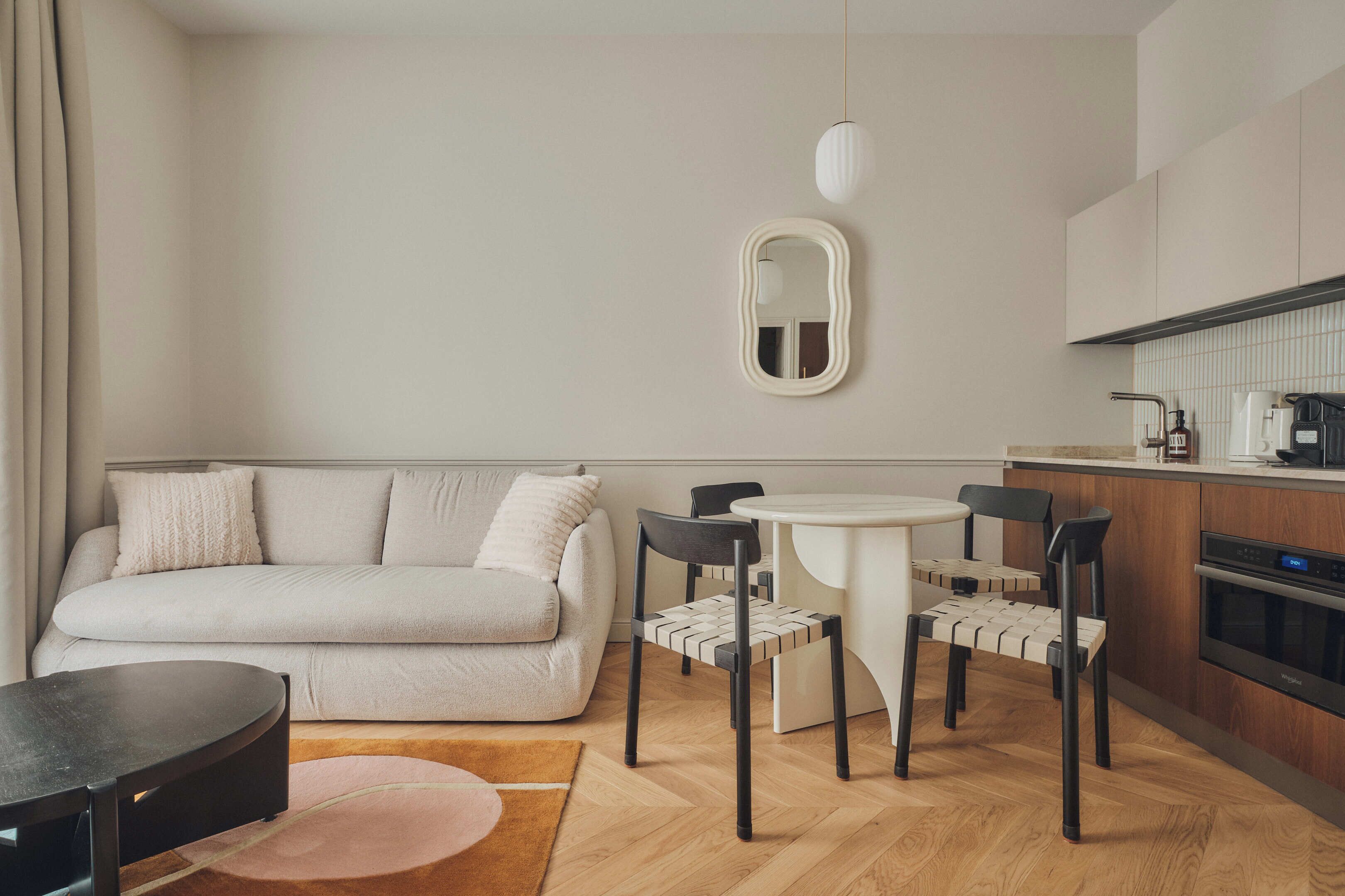 Una sala moderna con un sofá, una mesa redonda y dos sillas. Se observa un área de cocina con estufa y lavaplatos.