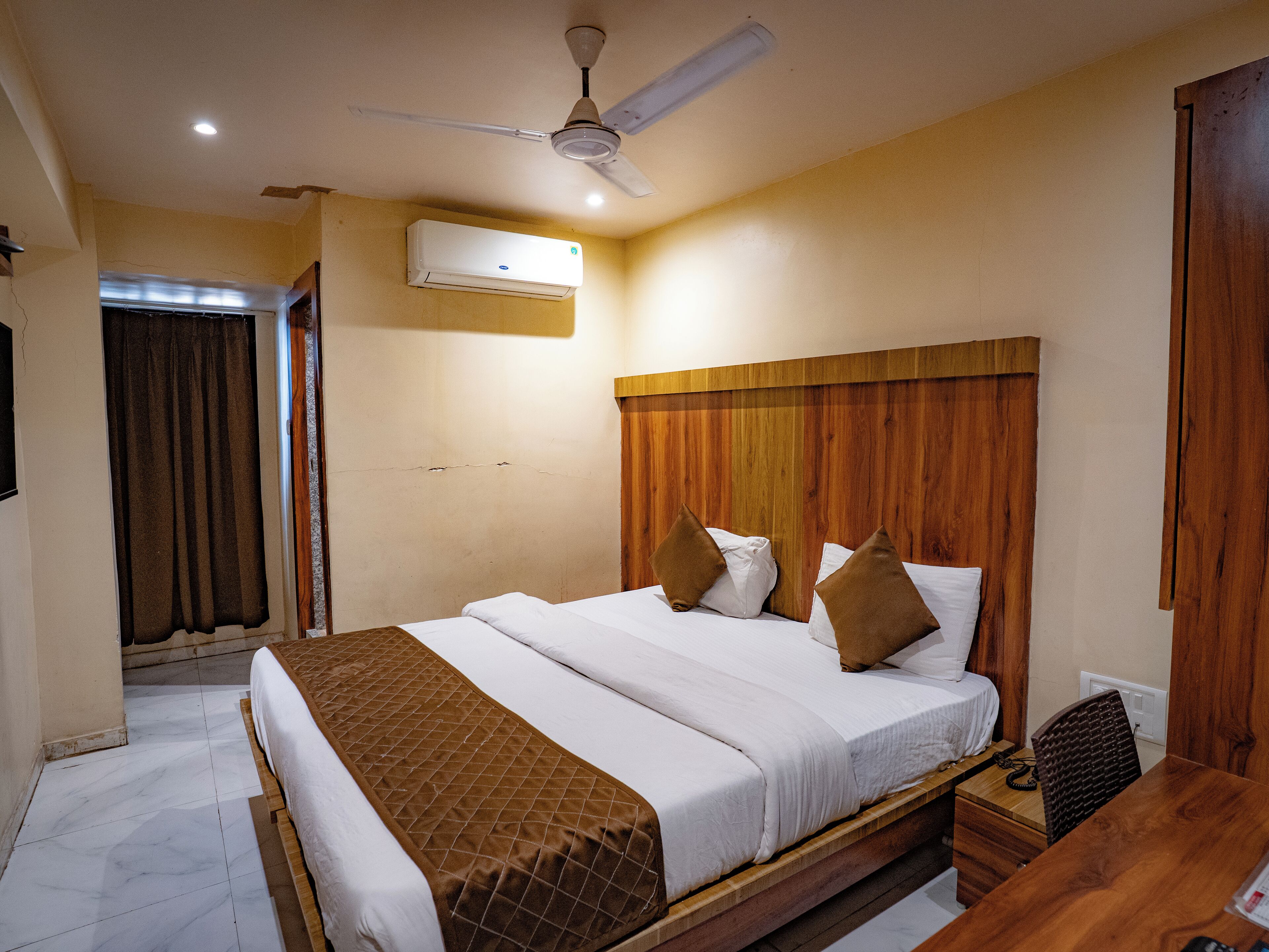 Een hotelkamer met een houten hoofdeinde, een bed met wit en bruin beddengoed, een plafondventilator, een airconditioning en een houten bureau met stoel.