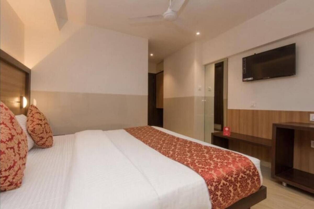 A hotel room with a bed, pillows, a TV mounted on the wall, and a wooden nightstand.