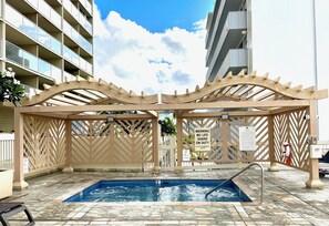 A swimming pool with a wooden pergola overhead, surrounded by stone tiles, and a lifeguard warning sign.