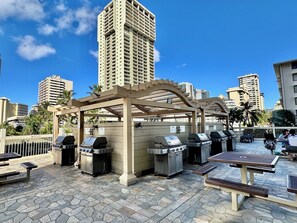 An outdoor patio area with barbecue grills and picnic tables, surrounded by high-rise buildings.