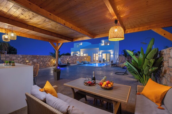 An outdoor patio with wicker furniture, a table with fruit and a bottle, and a pool in the background. - Amora Pool Villas - Villa 1 (XWX3+HGR Notia Rodos, Greece)