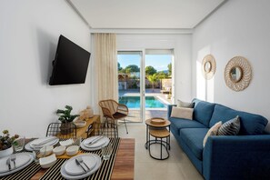 A modern living room with a blue sofa, a flat-screen TV, a dining area with a table set for four, and a view of a swimming pool through a glass door. - Amora Pool Villas - Villa 1 (XWX3+HGR Notia Rodos, Greece)